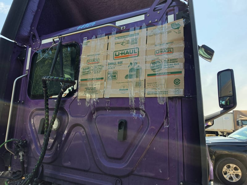 Driver-side door window missing from a semi truck cab, the opening temporarily covered with taped cardboard